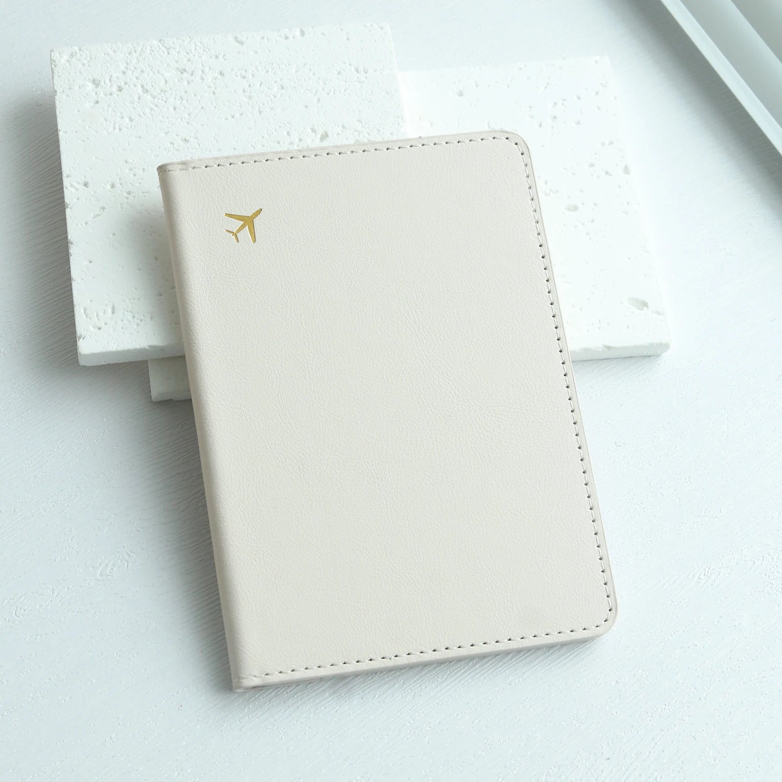 Minimalist beige passport holder with gold airplane icon on white textured background, Elarose