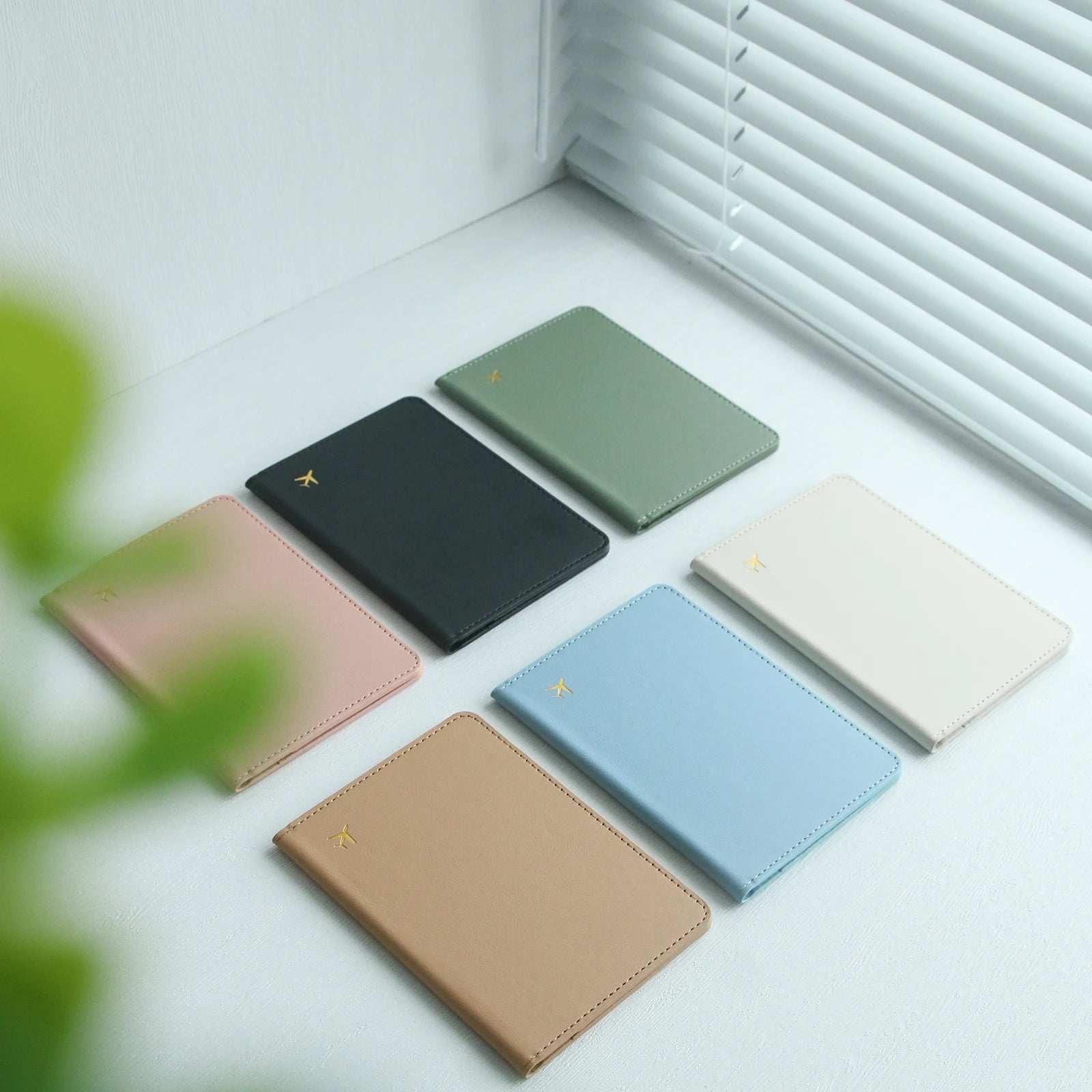 Assorted pastel and neutral Elarose passport holders on white desk by window blinds